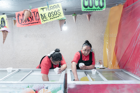 Two Hispanic adult women are serving ice creams on a cup in a street market stall
