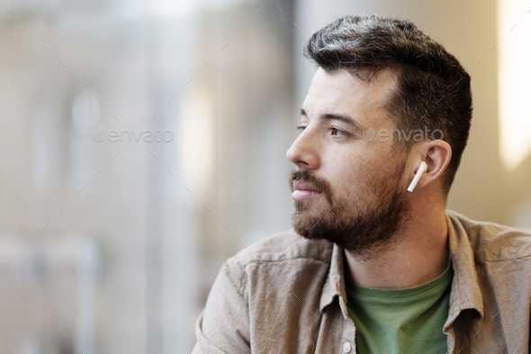 Pensive latin man listening music in modern earphones looking at window, copy space. Technology