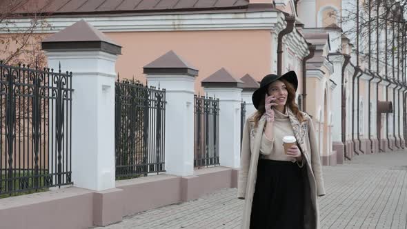 Young Redhaired Woman in a Coat and a Black Hat on the Background of the City Drinks Coffee and alt