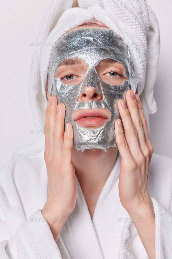 Vertical shot of serious young man applies silver sheet mask for reducing fine lines and skin