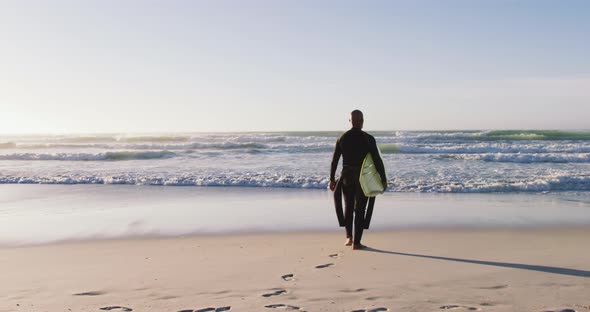 Senior african american man walking with a surfboard at the beach alt