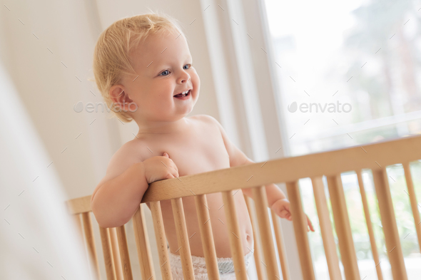 Indoor portrait of sweet little baby boy in diaper standing in bed trying to walk