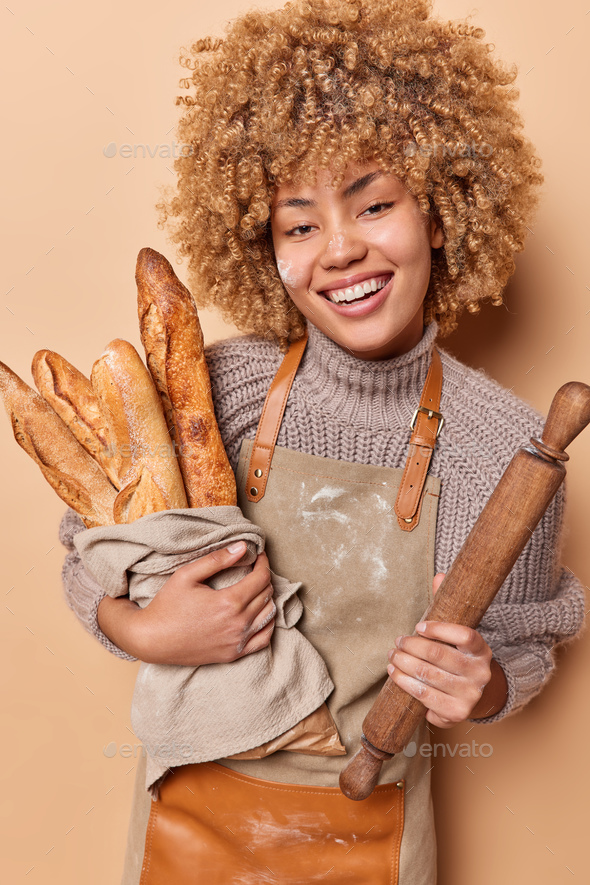 Positive busy curly female baker works in bakery shop baked delicious fresh baguettes holds wooden