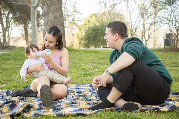 young couple sitting on the lawn while the woman feeds her baby a baby\'s bottle