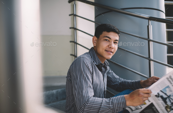 Asian millennial guy reading newspaper on stairs