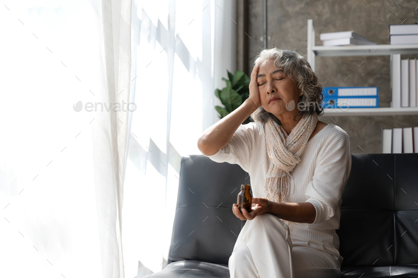 Depressed Asian senior woman sit on sofa looking away by the window. Loneliness elderly female