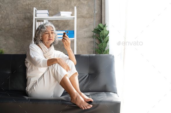 Depressed Asian senior woman sit on sofa looking away by the window. Loneliness elderly female