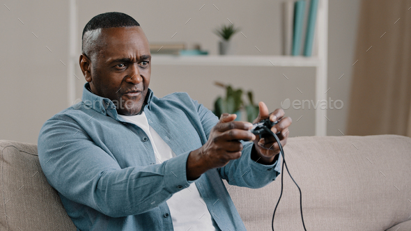 Focused mature african american man sitting in room on couch holding controller playing computer
