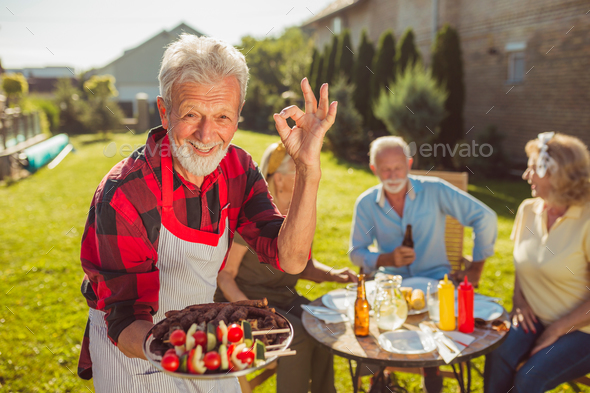 Senior man bringing food on a tray and showing OK sign while having outdoor lunch with friends