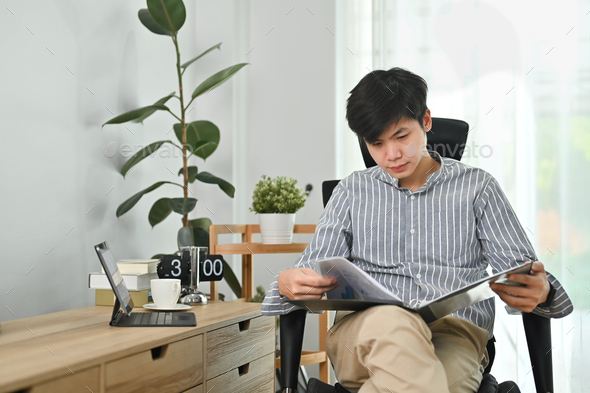 Successful start up businessman sitting on comfortable office chair, reading document in binder.