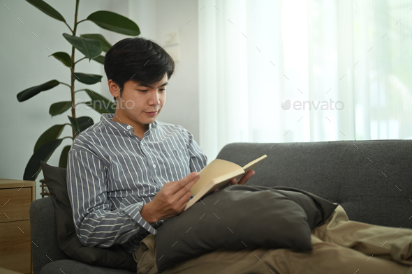 Calm millennial asian man relaxing co couch and reading literature story book in bright living room