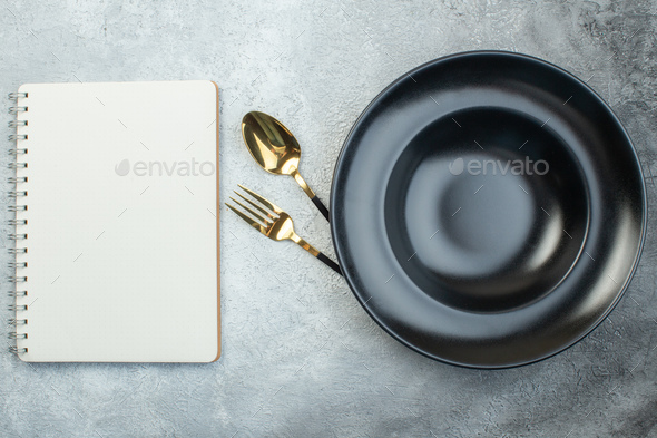Elegant cutlery set under black dinnerware set and spiral notebook on isolated gray ice background