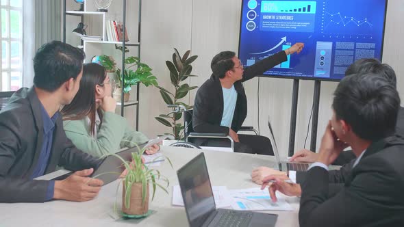 Businessman Sitting On Wheelchair Presentation To Business Colleagues, Pointing At Charts On TV alt