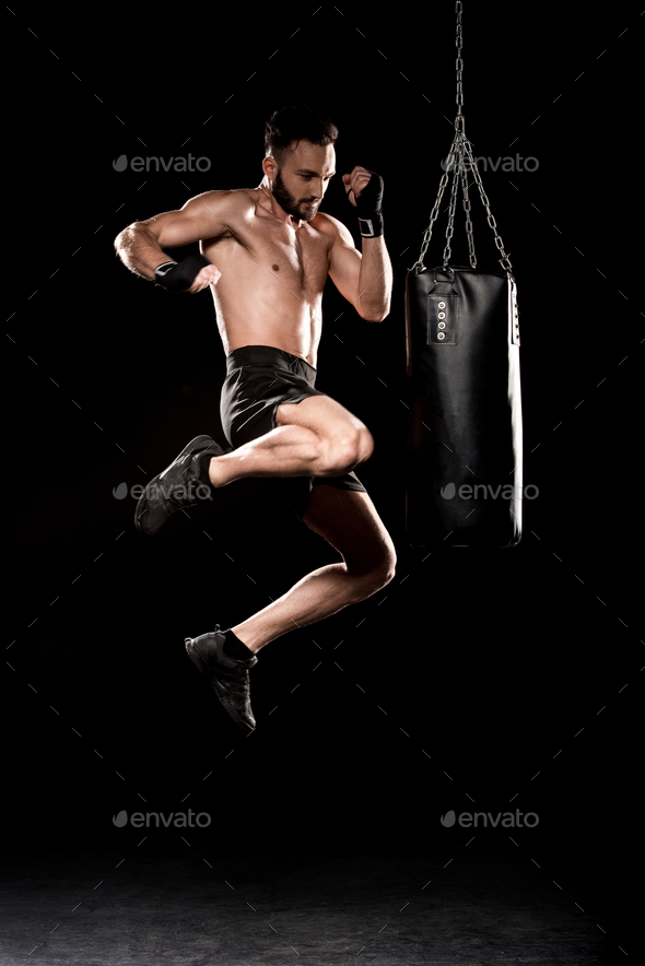 handsome boxer performing flying kick near punching bag on black background