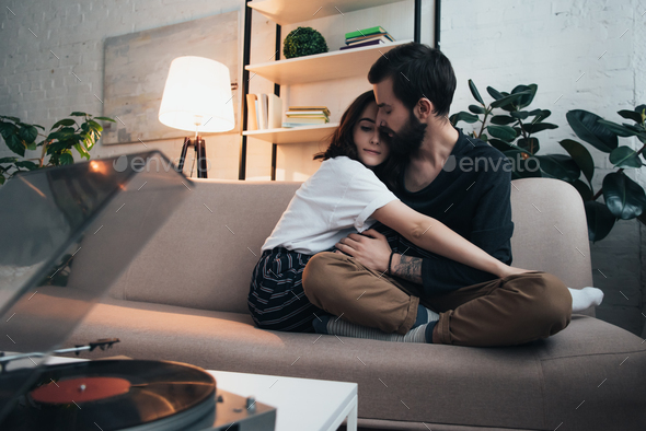 beautiful young couple sitting on couch, hugging and listening to vinyl record in living room
