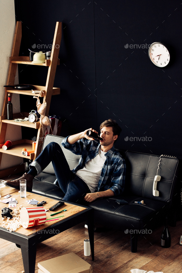 handsome man sitting on sofa and drinking beer at messy apartment
