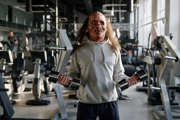 Zombie man with creepy face exercising with dumbbells at modern sport club