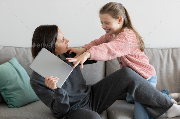 Strict european millennial mom takes laptop from angry little girl, quarrel in living room interior