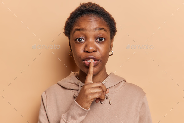 Keep silence please. Mysterious dark skinned young woman makes hush gesture looks wondered at camera