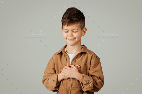 Positive cute little european child 6 years old in casual with closed eyes puts hands to chest