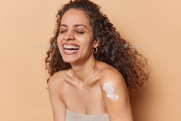 Body care. Cheerful curly haired woman keeps eyes closed applies moisturising lotion cream on arm