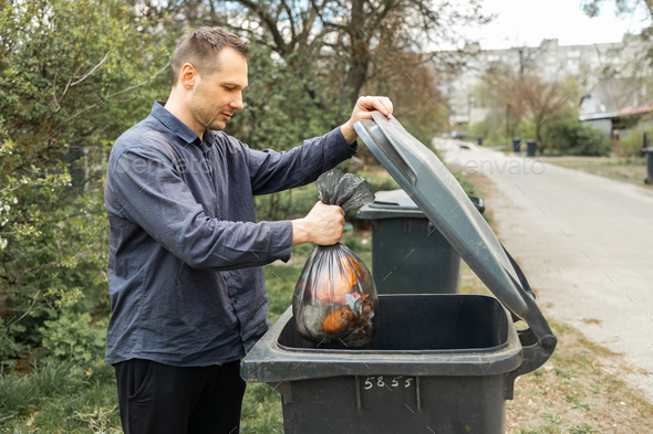 Man throwing out black eco-friendly recyclable trash bag in to big plastic green garbage container.