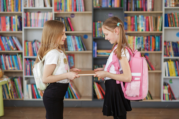 Two girls with backpacks meet and exchange books in school library. Benefits of everyday reading.