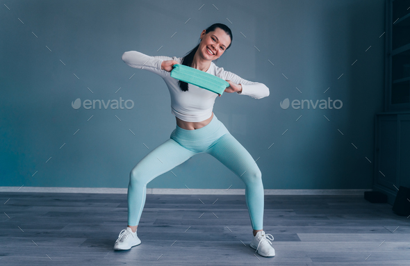 Brunette hispanic young woman in sportswear stretching rubber tape at fitness club looks at camera