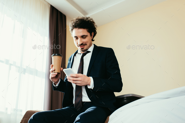 happy businessman using smartphone and holding disposable cup of coffee in hotel room