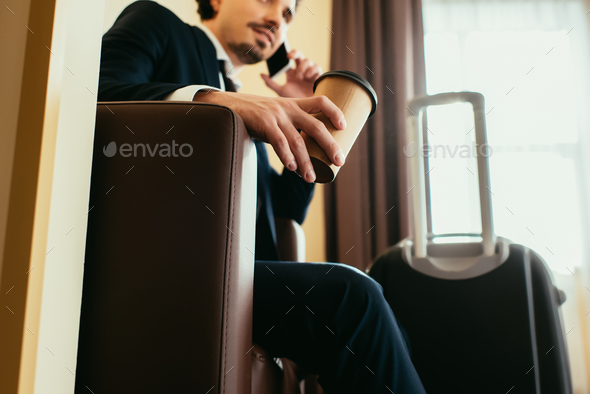 businessman with disposable cup of coffee talking on smartphone in hotel room with suitcase
