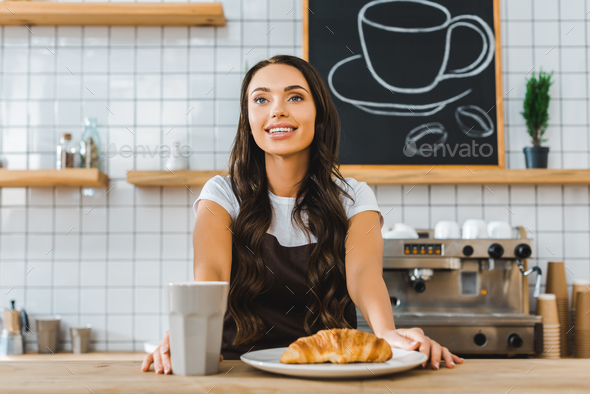 attractive cashier standing behind bar counter with cup, plate and croissant in coffee house