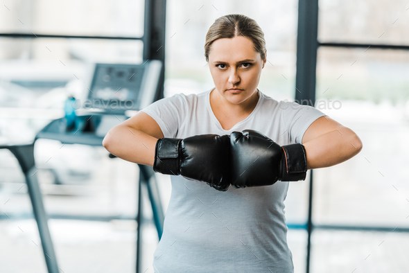 serious plus size woman wearing boxing gloves looking at camera in gym