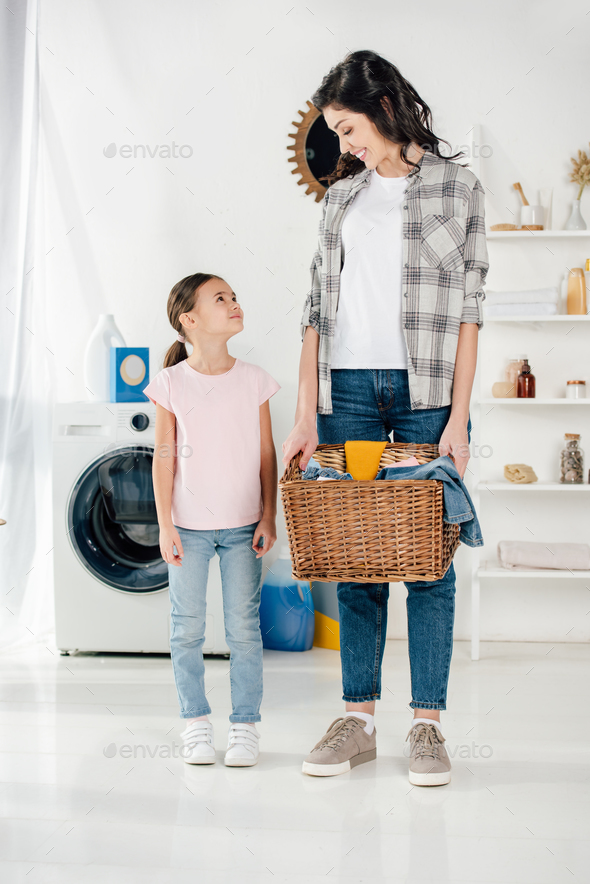 daughter in pink t-shirt standing near mother in grey shirt with basket and in laundry room