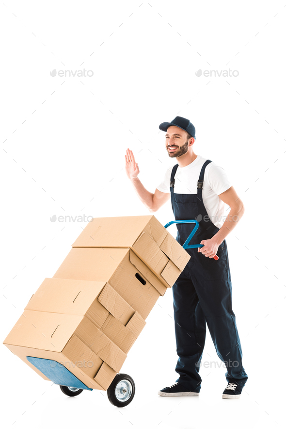 smiling delivery man transporting hand truck with cardboard boxes and showing hello gesture isolated