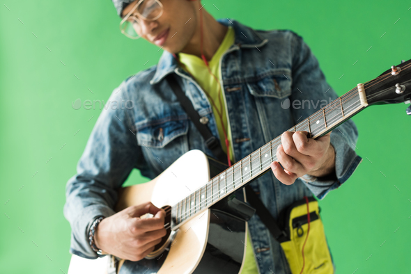 selective focus of mixed race man in denim playing acoustic guitar on green screen