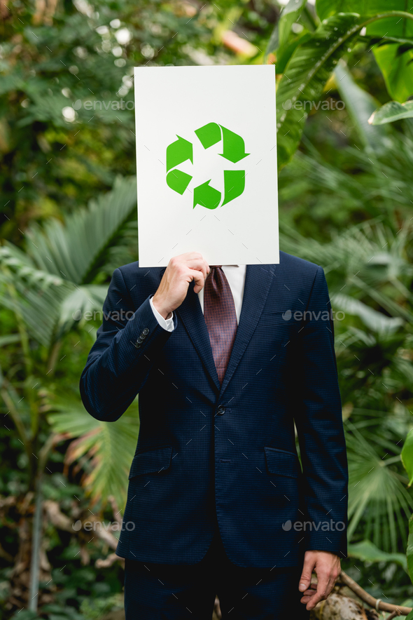 businessman holding card with green recycling sign in front of face in greenhouse
