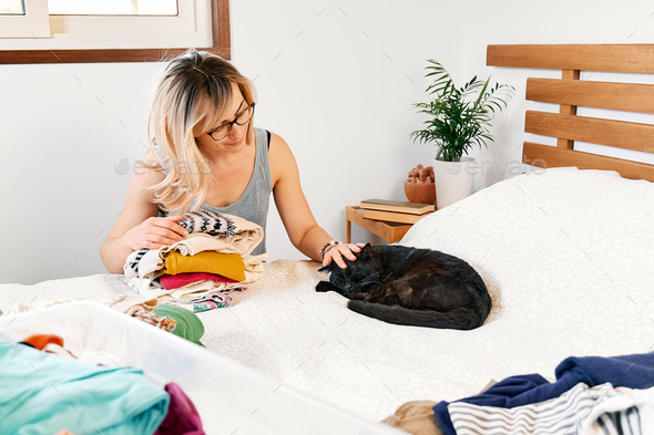 Blond woman making order of clothes arranging garments on bed and playing with her cat in bedroom.