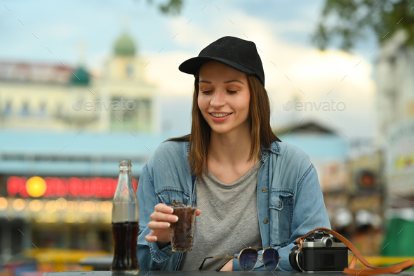 Hipster caucasian woman in hat drinking cola while sitting at street market