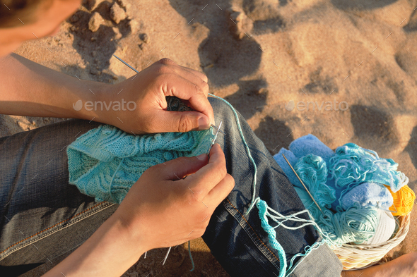 man knits blue yarn openwork pattern with metal knittings on beach