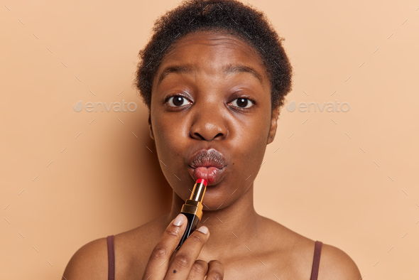 Close up shot of dark skinned woman with short curly hair applies lipstick puts on everyday makeup