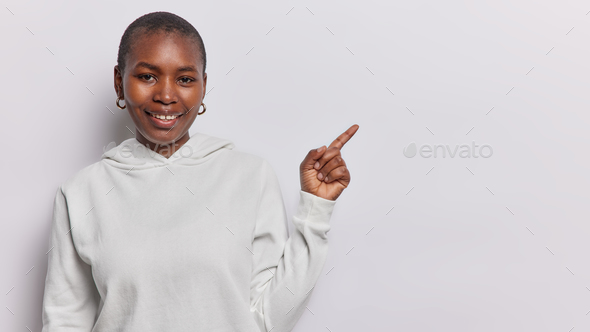 Pleased dark skinned curly haired Afro American woman dressed in sweatshirt points index finger on