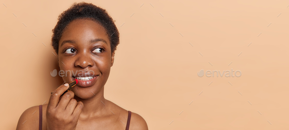 Beauty portrait of Afro American woman with short hair applies lipstick looks aside puts on organic