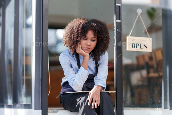 Female shopkeeper sitting stressed out at the store entrance frustrated by the economic impact.