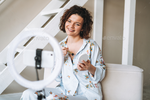 Young woman blogger in home clothes doing makeup with mobile phone and ring lamp online in studio - Stock Photo - Images