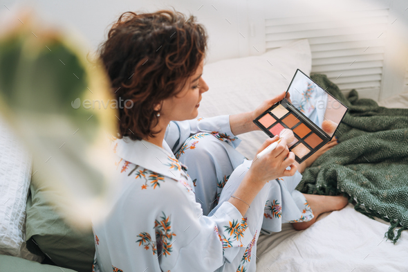 Young woman in blue pajamas applies makeup with face sculpting palette sitting on bed at home - Stock Photo - Images
