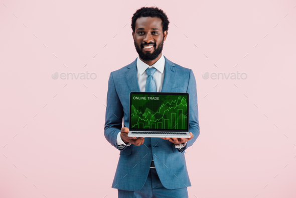 smiling african american businessman showing laptop with online trade isolated on blue