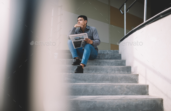 Asian millennial guy reading newspaper on stairs