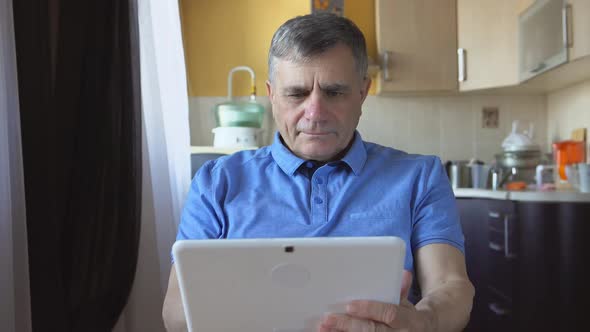 Potrait Of An Aged Male At The Blue Shirt Sits And Uses A White Tablet Pc At Home