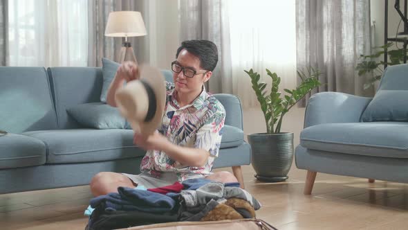 Asian Young Man Try Hat And Packing Clothes In Suitcase At Home, Preparing For Vacation alt