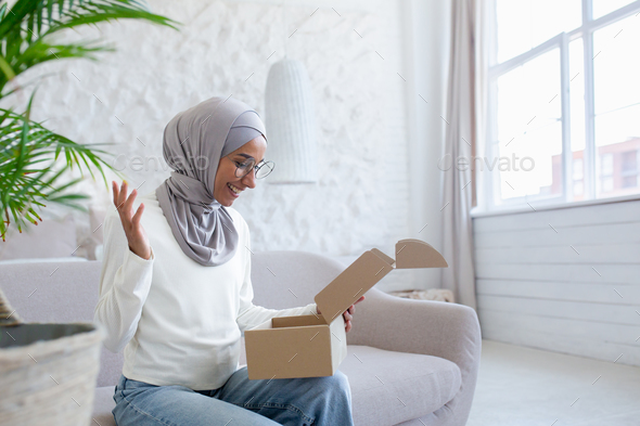 Home delivery of mail. Young Muslim woman in hijab received parcel, happy sitting on sofa and